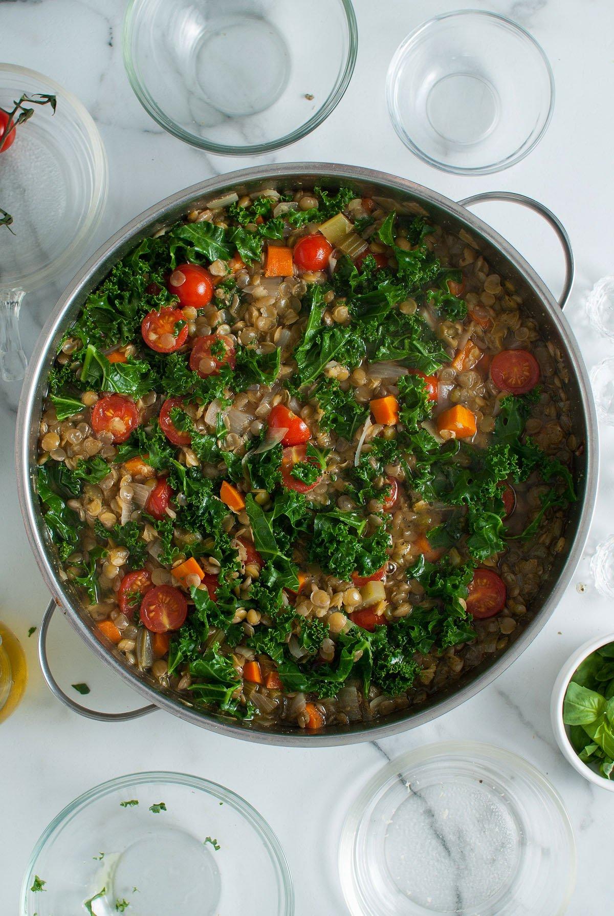Μια κατσαρόλα Zuppa Di Lenticchie, μια ιταλική σούπα φακής που ξεχειλίζει από φρέσκα λαχανικά όπως λάχανο κέιλ, ντοματίνια, φακές και καρότα, βρίσκεται περιτριγυρισμένη από άδεια γυάλινα μπολ.