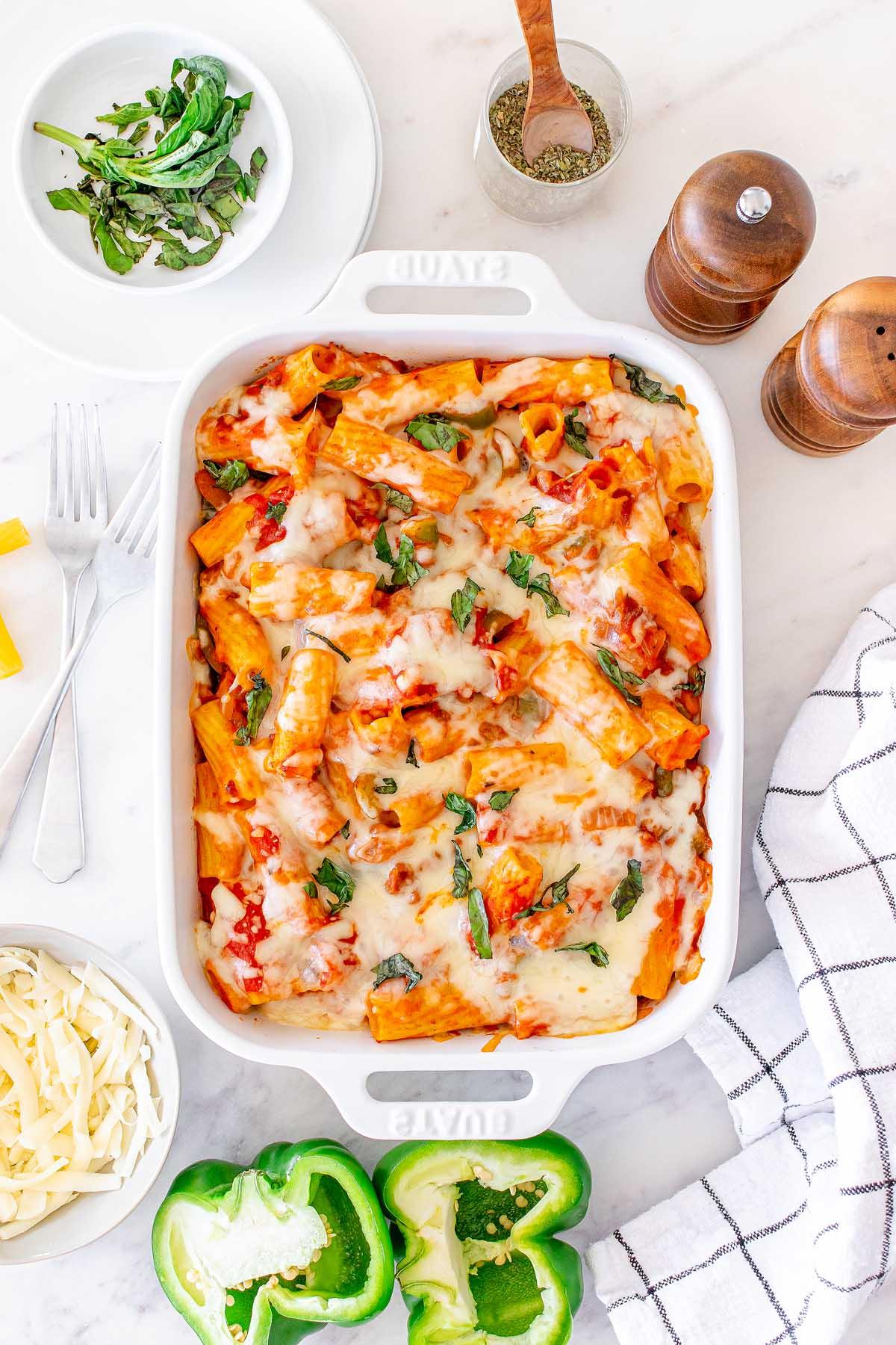 Ένα ψημένο Pasta al Forno με λιωμένο τυρί και βασιλικό σε ένα λευκό πυρίμαχο σκεύος, περιτριγυρισμένο από πράσινες πιπεριές, τριμμένο τυρί, μαχαιροπίρουνα και μπαχαρικά σε έναν μαρμάρινο πάγκο.