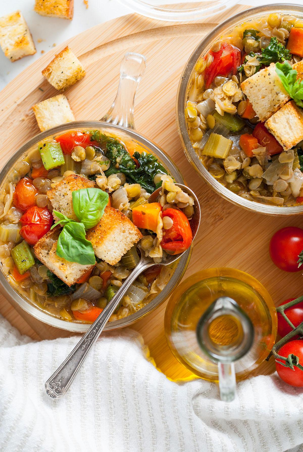 Δύο μπολ Zuppa Di Lenticchie, μια χορταστική μινεστρόνε φακής που ξεχειλίζει από φρέσκα λαχανικά, γαρνιρισμένα με κρουτόν και βασιλικό, με ντομάτες και ελαιόλαδο κοντά.