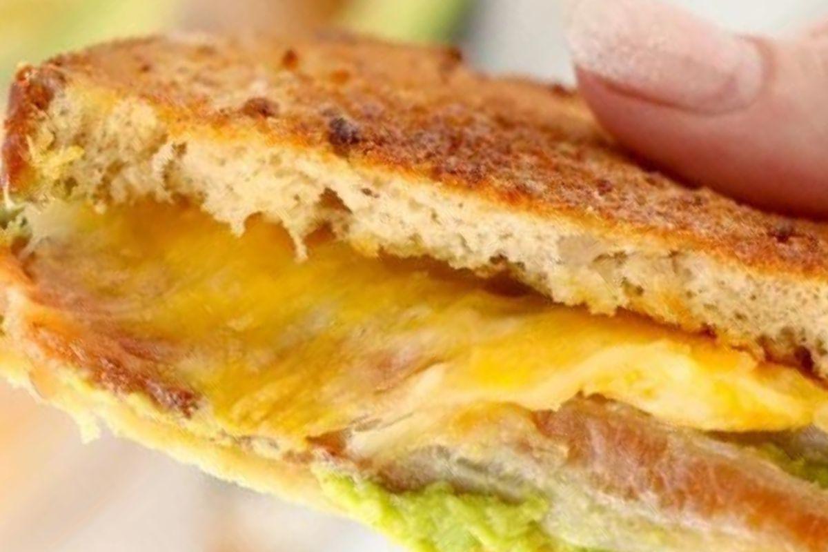 Κοντινό πλάνο συνταγών grilled cheese που δείχνουν ένα σάντουιτς με ένα κομμάτι που λείπει, αποκαλύπτοντας λιωμένο τυρί και αβοκάντο στο εσωτερικό.
