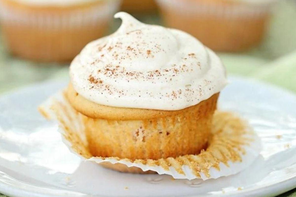 Ένα cupcake βανίλιας με γλάσο καραμέλας και μια πρέζα κανέλα από πάνω, ξετυλιγμένο και τοποθετημένο σε ένα λευκό πιάτο.