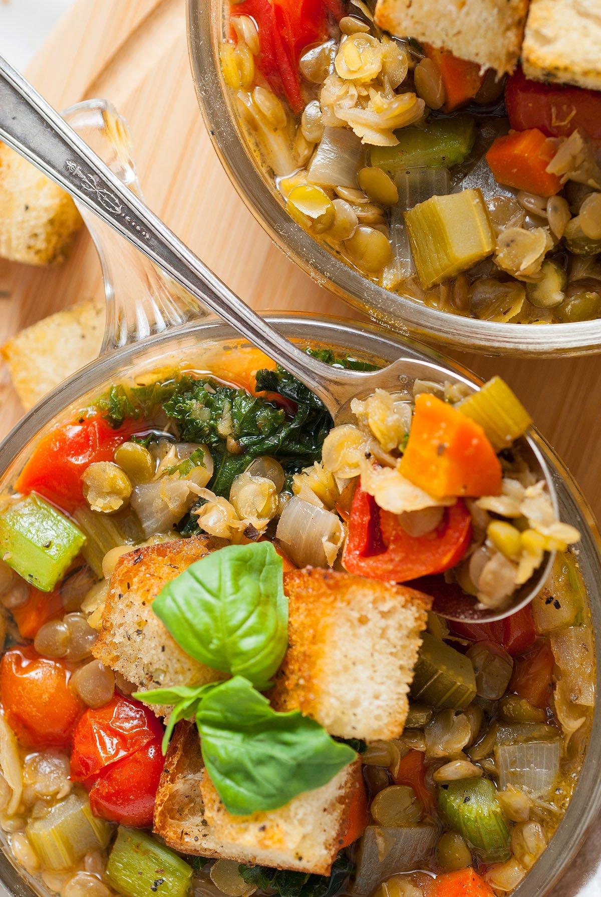 Δύο μπολ Zuppa Di Lenticchie, μια χορταστική ιταλική σούπα λαχανικών με κρουτόν και φρέσκο βασιλικό, σερβιρισμένα με μεταλλικά κουτάλια.