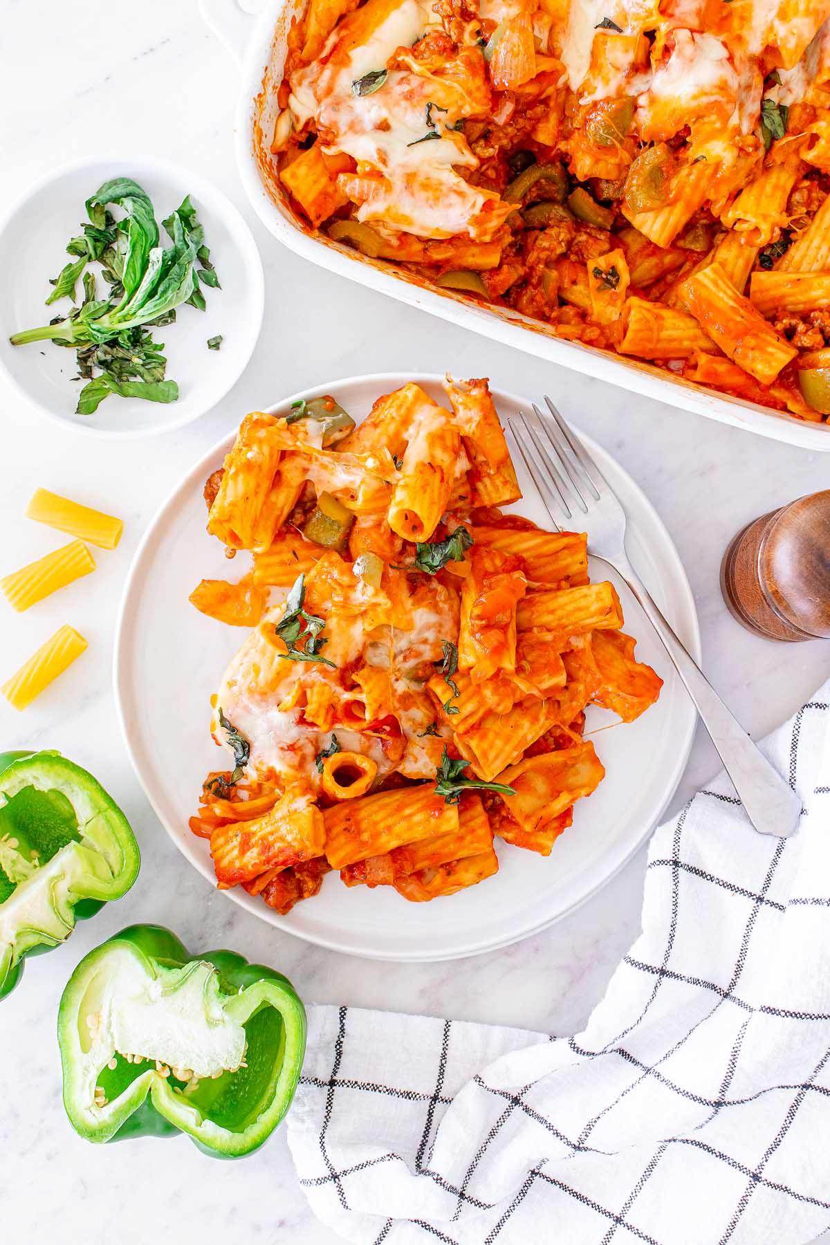 Ένα πιάτο Pasta al Forno με ψητά ριγκατόνι και λιωμένο τυρί, σερβιρισμένο με φρέσκο βασιλικό και πράσινες πιπεριές, δίπλα σε ένα λευκό μπολ που περιέχει επιπλέον βασιλικό και μισή πράσινη πιπεριά.