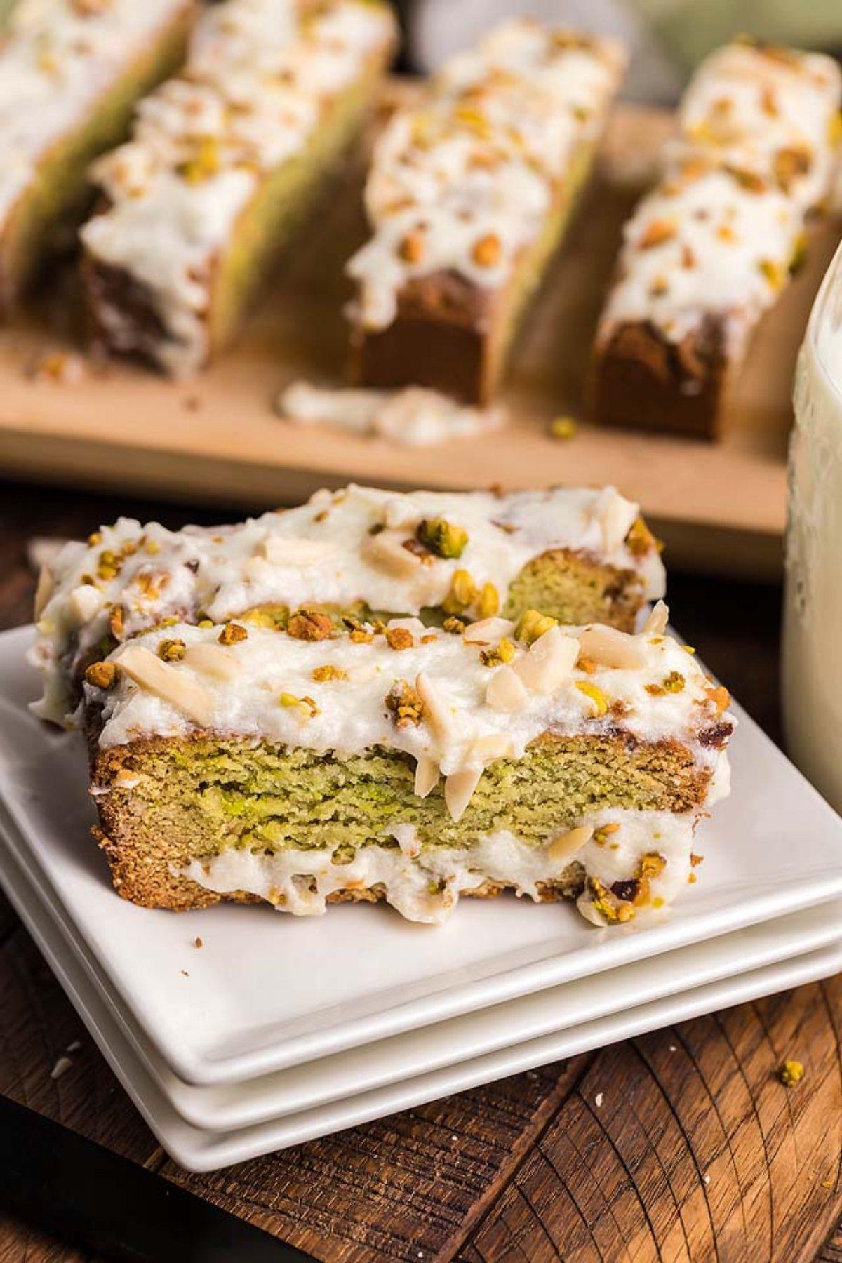 Μια φέτα Air Fryer Pistachio Coffee Cake με λευκό γλάσο βρίσκεται σε στοιβαγμένα πιάτα, με περισσότερες νόστιμες φέτες να παρατάσσονται στο παρασκήνιο.