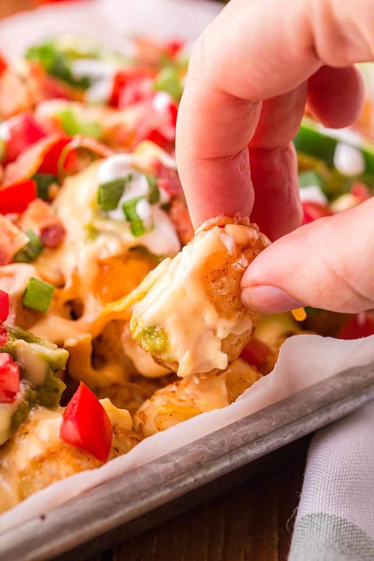 Ένα χέρι σηκώνει ένα τυρένιο, φορτωμένο tater tot nacho με ξινή κρέμα, ψιλοκομμένες ντομάτες και φρέσκα κρεμμυδάκια από ένα ταψί.