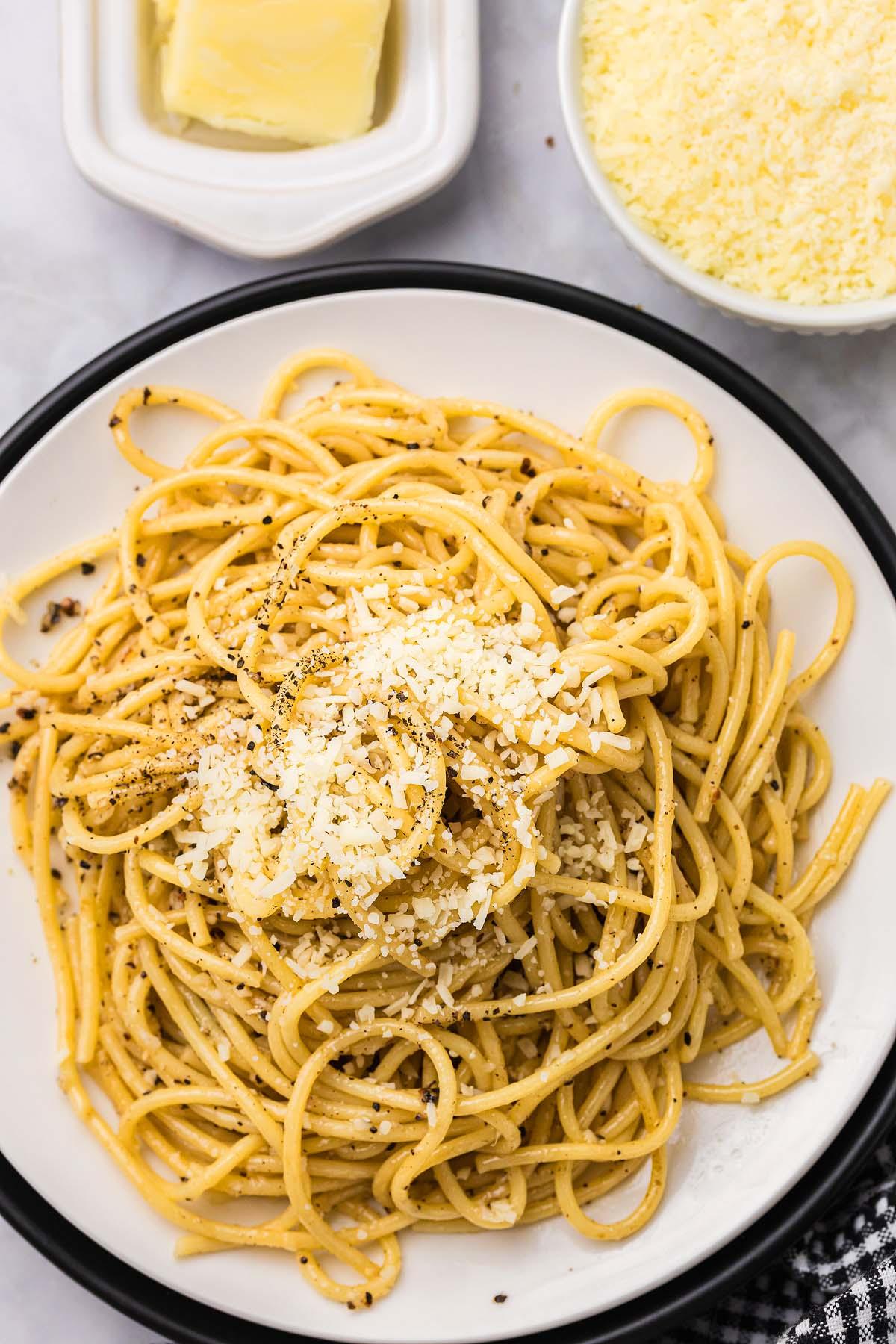 Ένα πιάτο cacio e pepe περιλαμβάνει σπαγγέτι καρυκευμένα με μαύρο πιπέρι και τριμμένο τυρί. Δίπλα στο πιάτο, ένα μπολ με τριμμένο τυρί και ένα κομμάτι βούτυρο ολοκληρώνουν τη σκηνή.