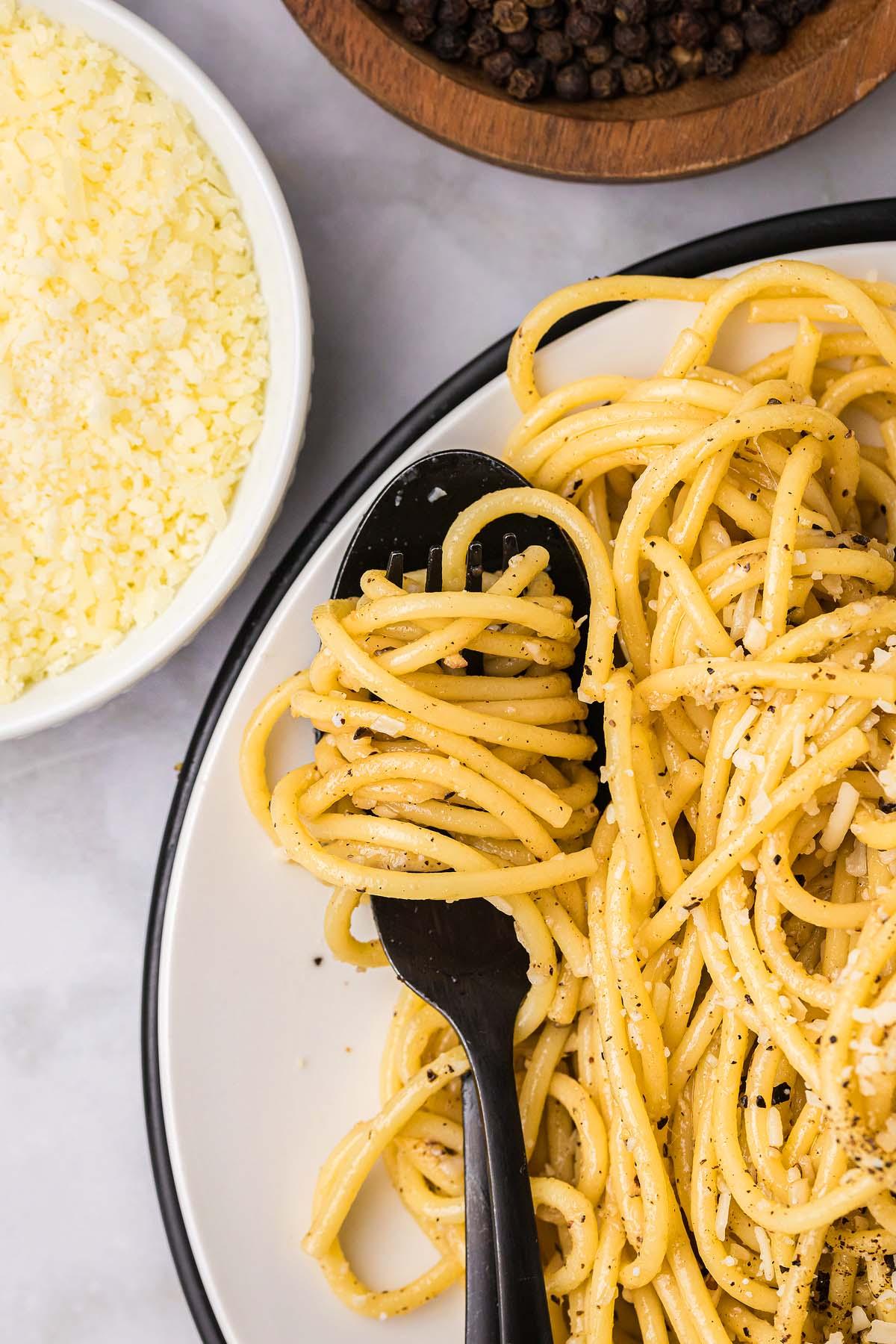 Ένα πιάτο cacio e pepe, καρυκευμένο με μαύρο πιπέρι και τυρί, περιλαμβάνει noodles κομψά τυλιγμένα γύρω από ένα μαύρο πιρούνι. Κοντά, ένα μπολ με τριμμένο τυρί και ολόκληρους κόκκους πιπεριού ενισχύουν τη μινιμαλιστική γοητεία αυτού του κλασικού πιάτου.