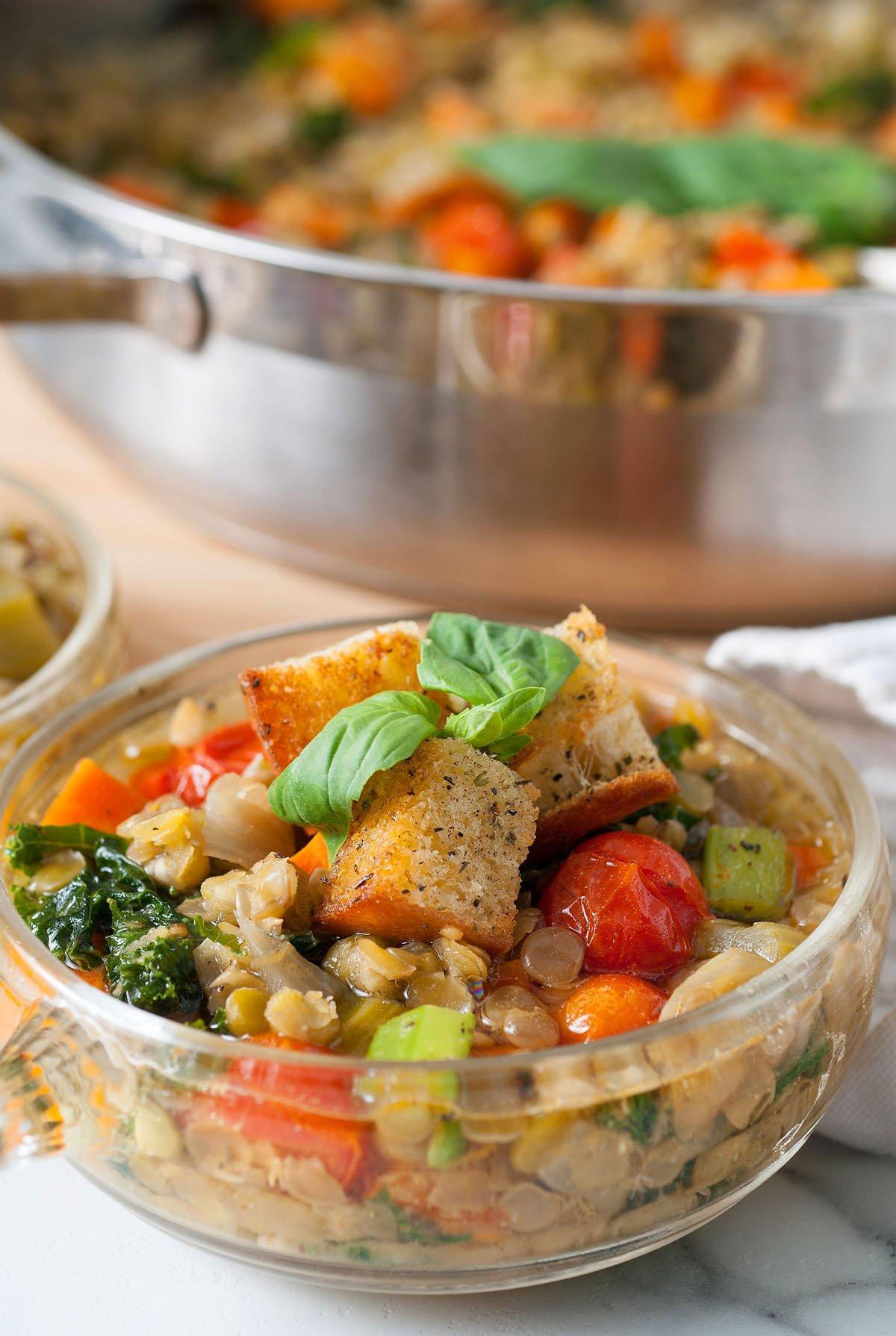Ένα μπολ με ιταλική σούπα φακής – Zuppa Di Lenticchie με κρουτόν και βασιλικό, συνοδευόμενο από μια κατσαρόλα με γευστική ιταλική σούπα λαχανικών στο φόντο.