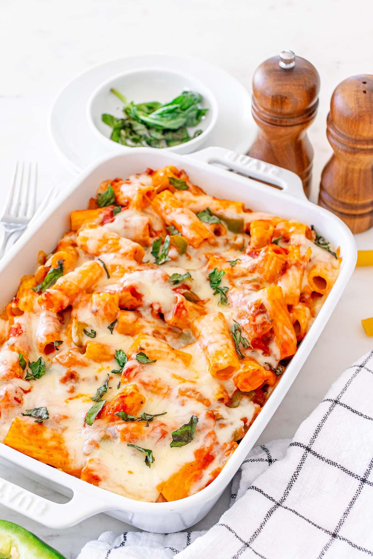 Ένα λευκό πυρίμαχο σκεύος γεμάτο με ιταλικό Pasta al Forno, με λιωμένο τυρί και βότανα. Δύο ξύλινες αλατοπιπεριέρες, ένα μικρό πιάτο με χόρτα και ένα καρό ύφασμα βρίσκονται κοντά.