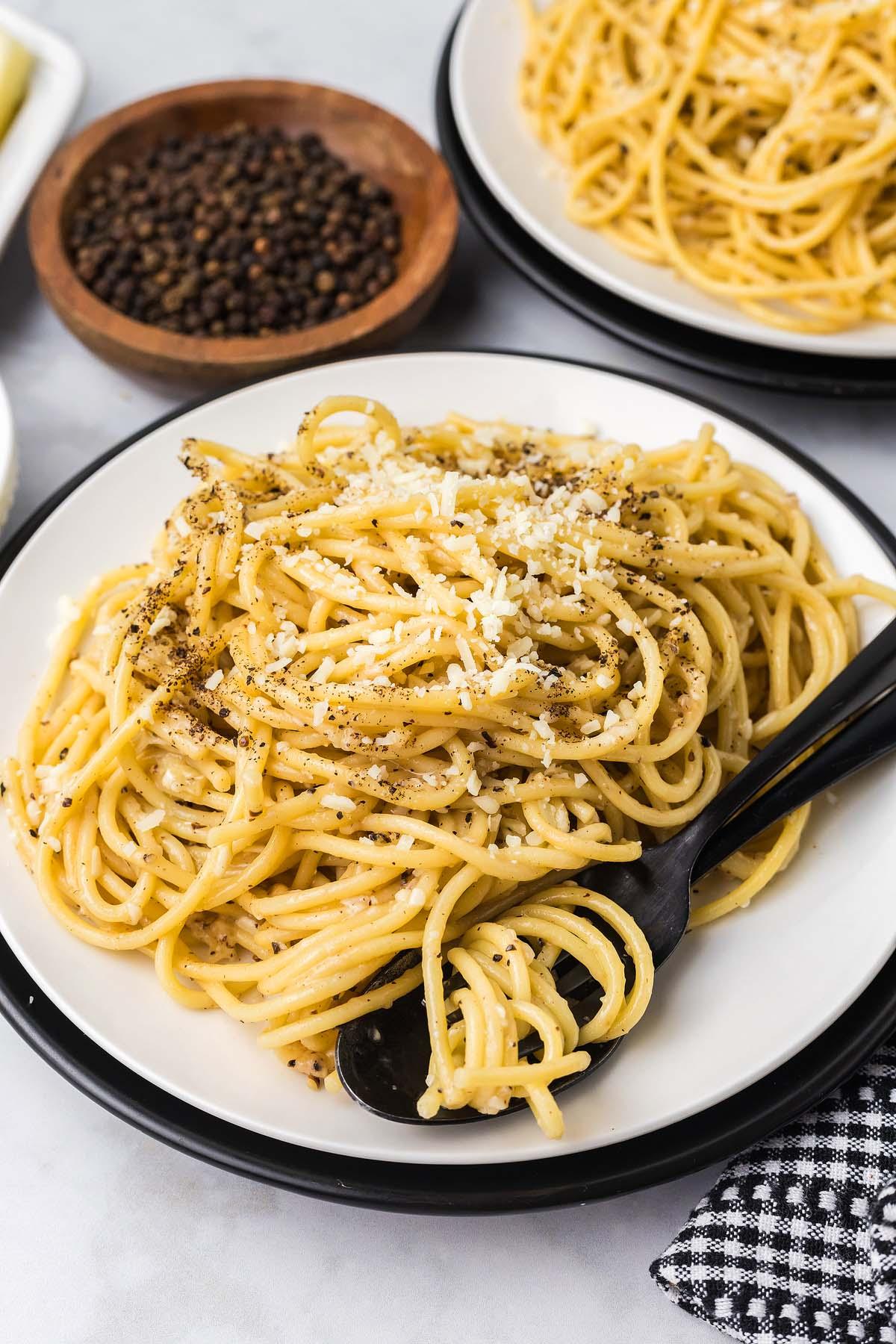 Ένα πιάτο cacio e pepe, με σπαγγέτι γαρνιρισμένα με τριμμένο τυρί και πιπέρι, κομψά σε συνδυασμό με ένα μαύρο πιρούνι. Επιπλέον πιάτα και μπολ γεμάτα πιπέρι βρίσκονται δελεαστικά στο βάθος.