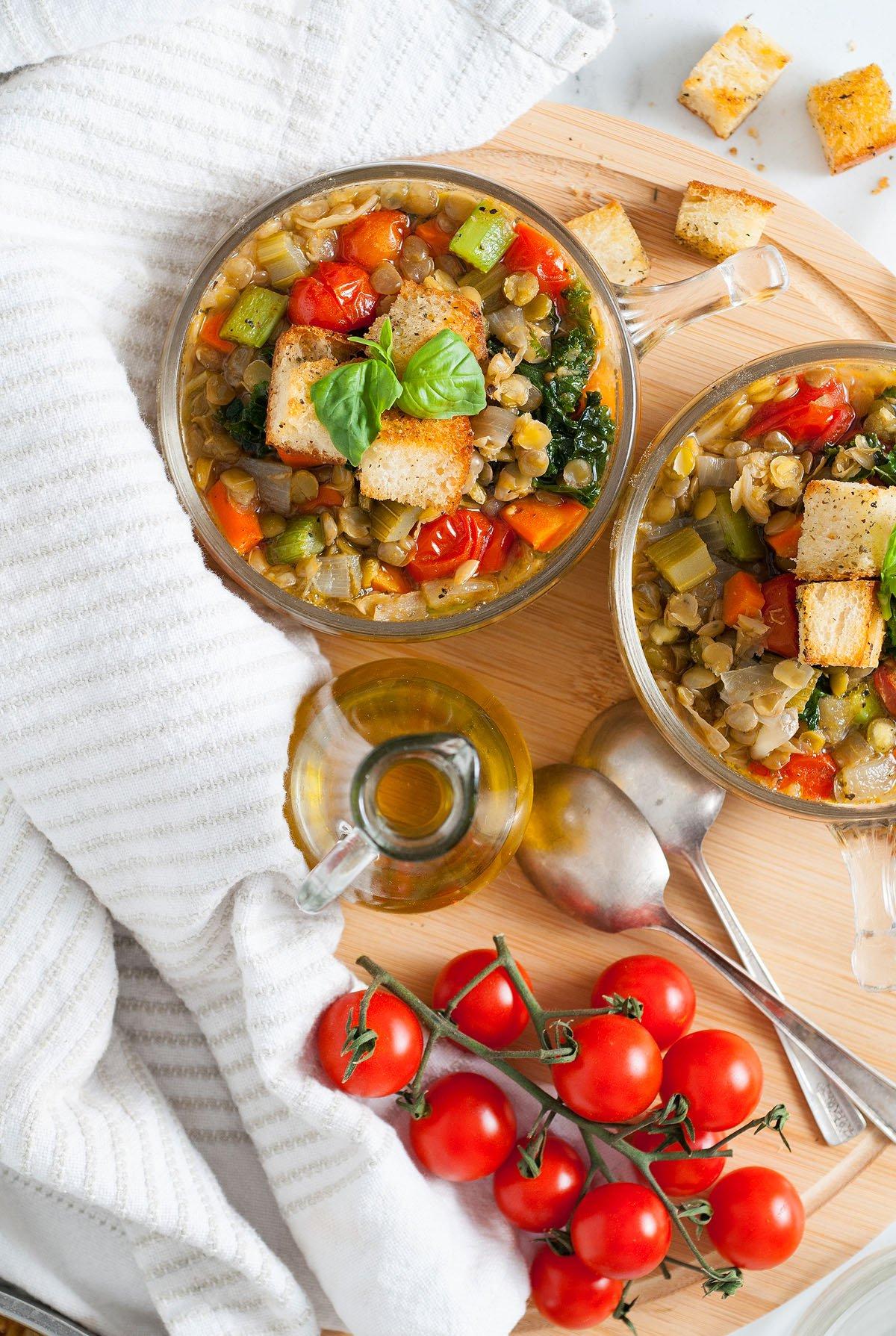 Δύο μπολ με ιταλική σούπα φακής – Zuppa Di Lenticchie με κρουτόν, βασιλικό και ντοματίνια ακουμπούν σε ένα ξύλινο ταμπλό δίπλα σε μια ριγέ πετσέτα.