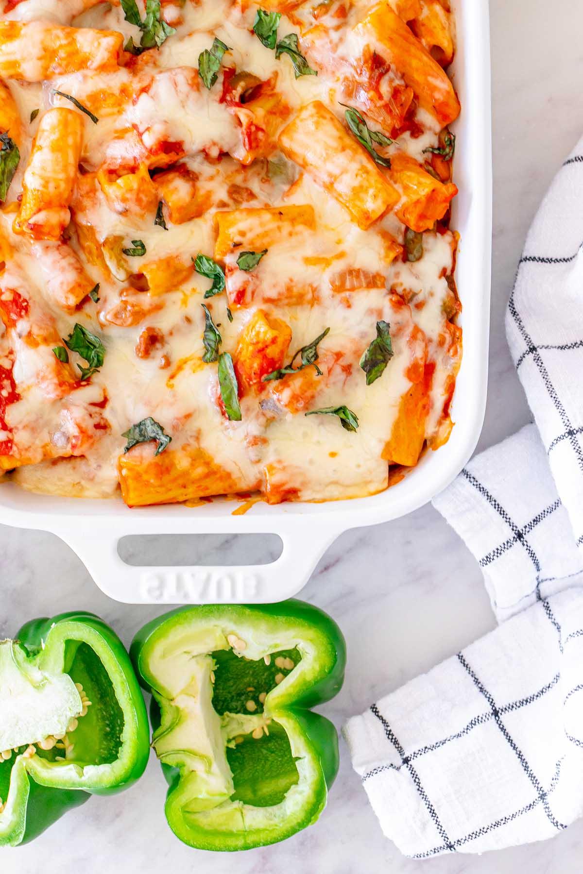 Αυθεντικό pasta al forno με λιωμένο τυρί και φρέσκα βότανα σε ένα λευκό ταψί δίπλα σε μια κομμένη πράσινη πιπεριά και ένα ριγέ ύφασμα.