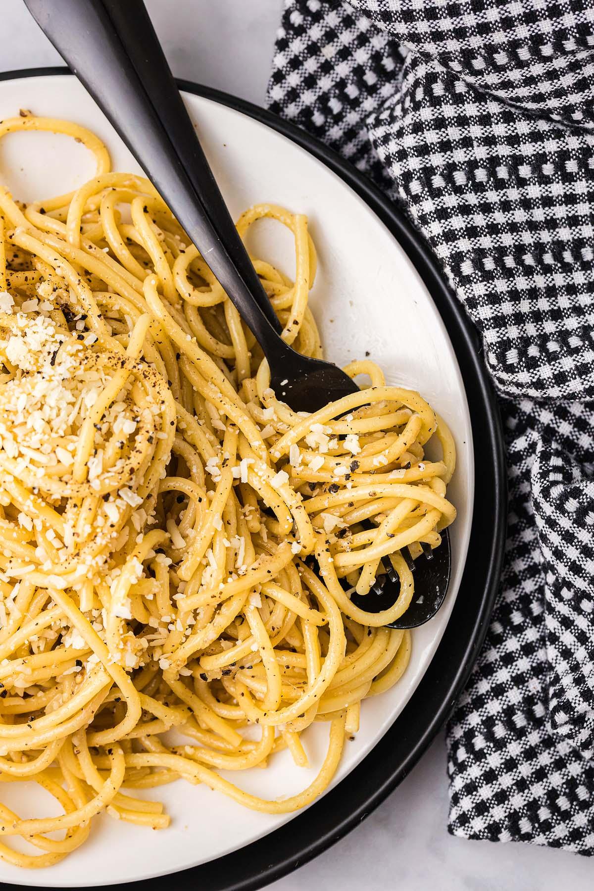 Cacio e pepe κομψά σερβιρισμένο σε ένα πιάτο με τυρί και μαύρο πιπέρι, συνοδευόμενο από ένα μαύρο πιρούνι που ακουμπά δίπλα του σε ένα ασπρόμαυρο καρό ύφασμα.