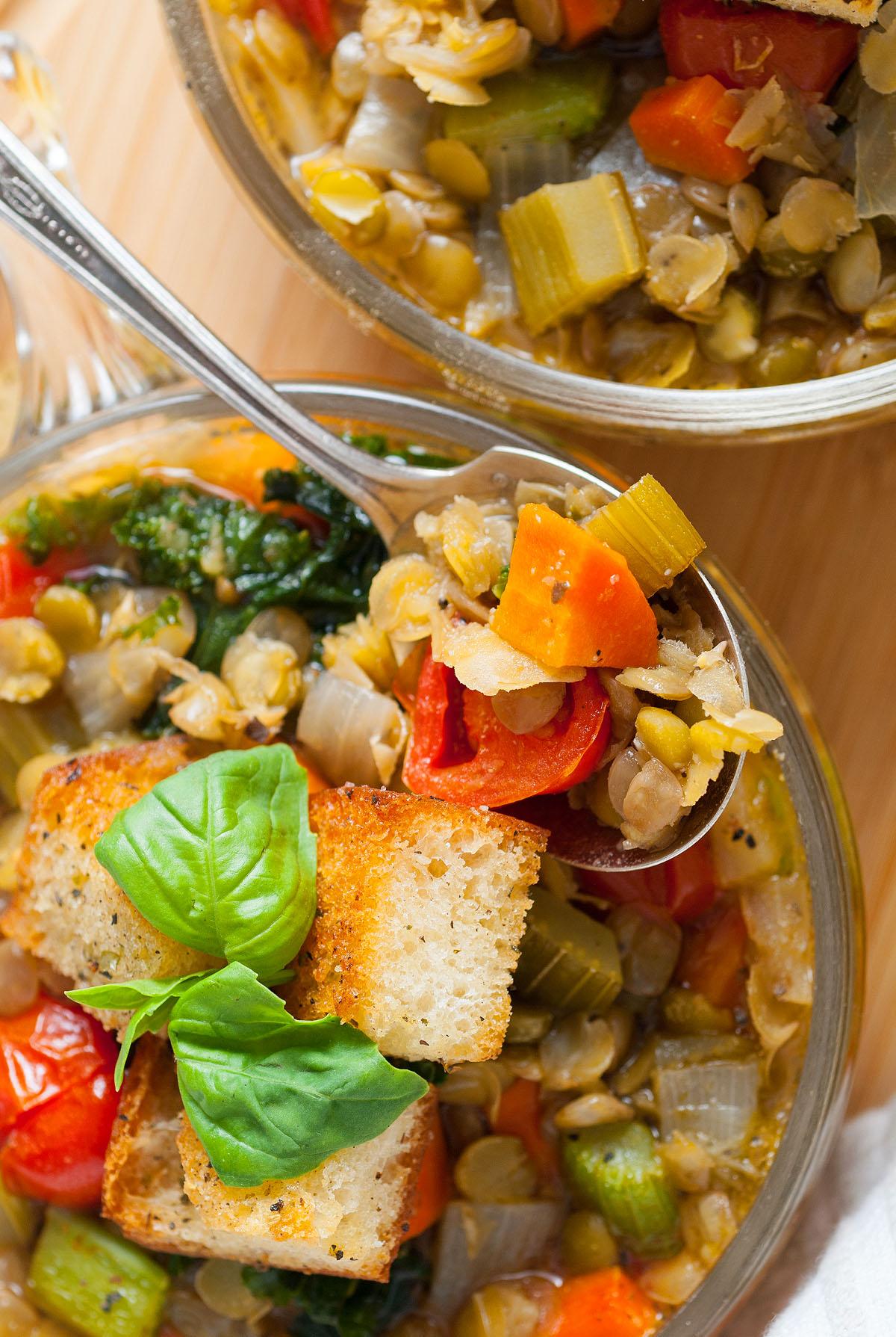 Ένα μπολ με ιταλική σούπα φακής – Zuppa Di Lenticchie, γεμάτο με φρέσκα λαχανικά και γαρνιρισμένο με κρουτόν και φύλλα βασιλικού, βρίσκεται δίπλα σε μια κουταλιά σούπας.