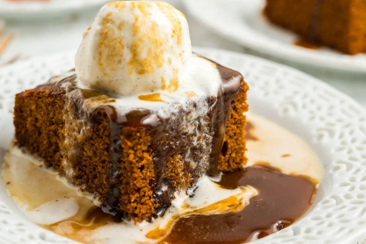 Μια φέτα sticky toffee pudding με μια μπάλα παγωτό βανίλια, σερβιρισμένη σε ένα λευκό πιάτο με καραμέλα.