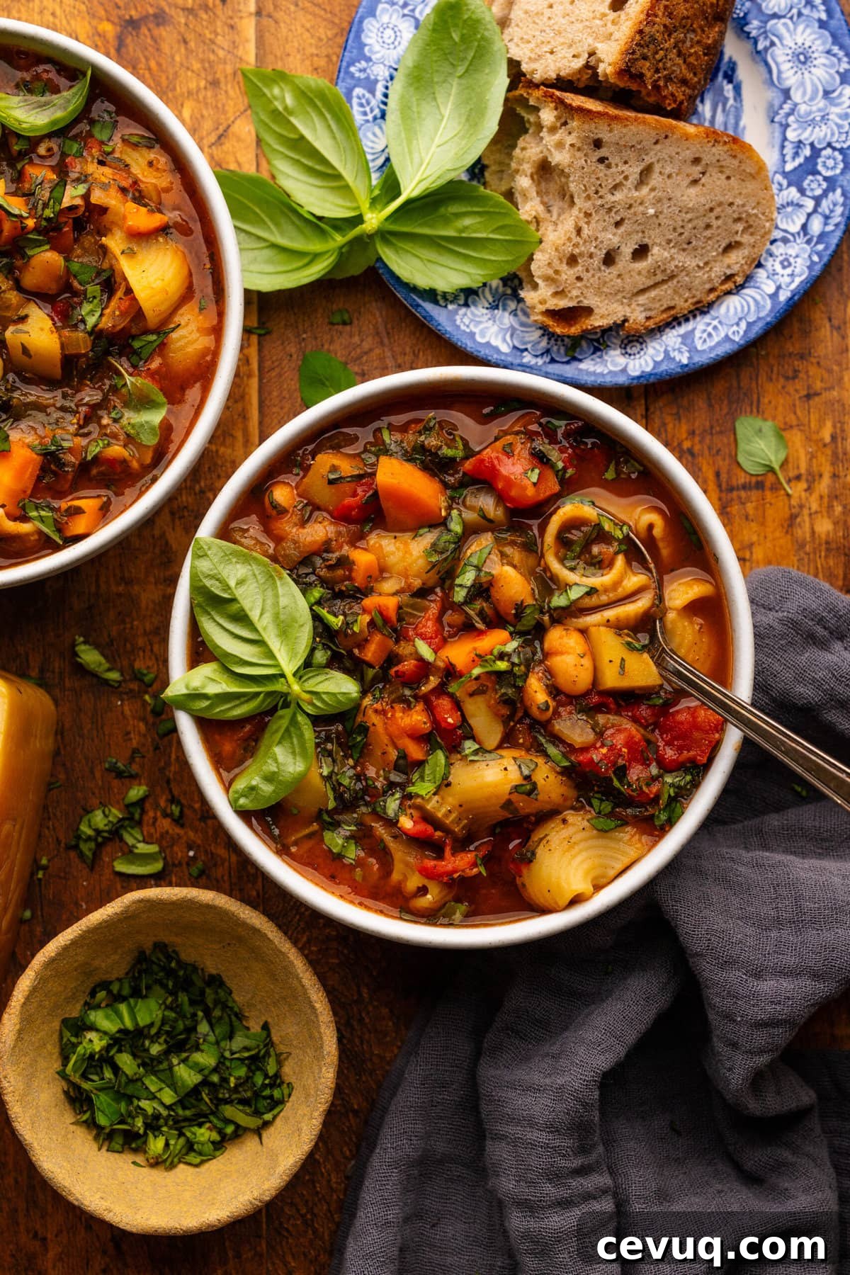 Two bowls with minestrone soup in them. The two bowls are displayed on a wooden background with bread on a plate, a small bowl of chopped herbs, and a gray napkin placed around the one bowl.