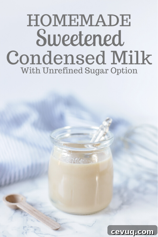 Homemade sweetened condensed milk in a glass jar with an antique spoon in it. A wooden spoon is sitting next to the jar and a blue and white striped towel in the back