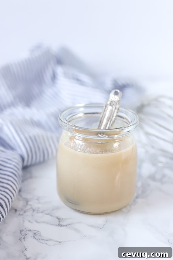 glass jar of homemade sweetened condensed milk with an antique silver spoon coming out. a blue and white striped towel is in the background