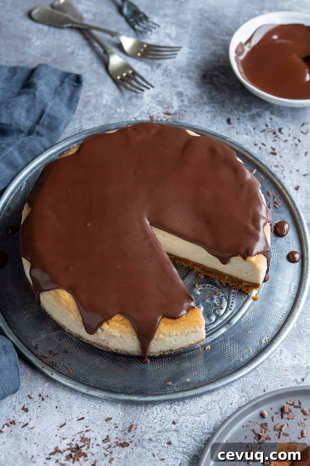 Vegan baileys cheesecake topped with chocolate ganache on a metal plate on a grey background with a bowl of ganache and three forks.