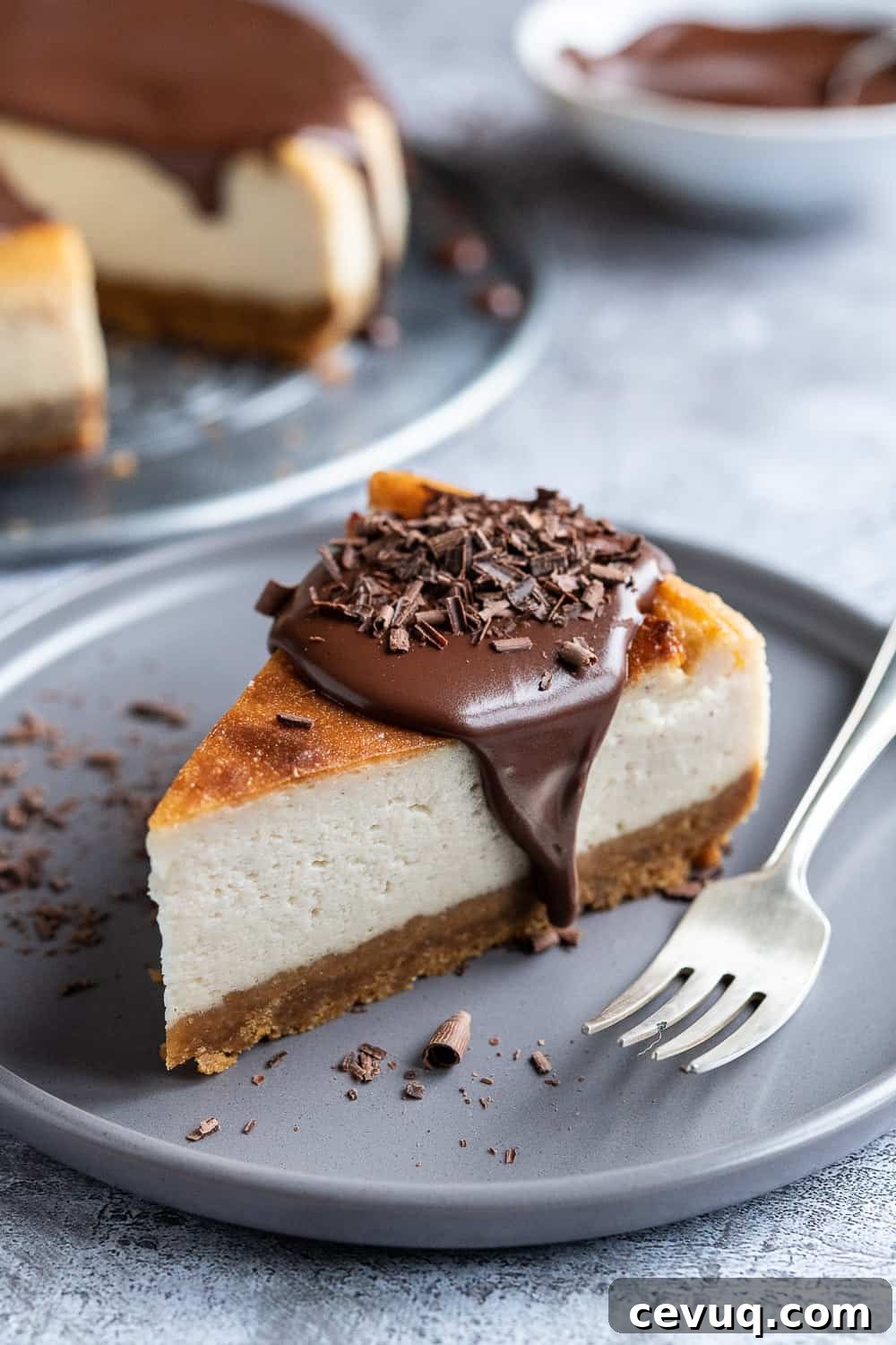 a slice of vegan baileys cheesecake topped with ganache and chocolate shavings on a grey plate with a silver fork.