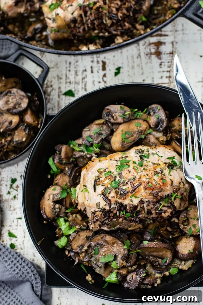 A dish with wild rice stuffed pork chops lay in a bed of mushrooms with a fork and knife sitting on the dish. A cast iron skill sits above the dish with a gray napkin next to it.
