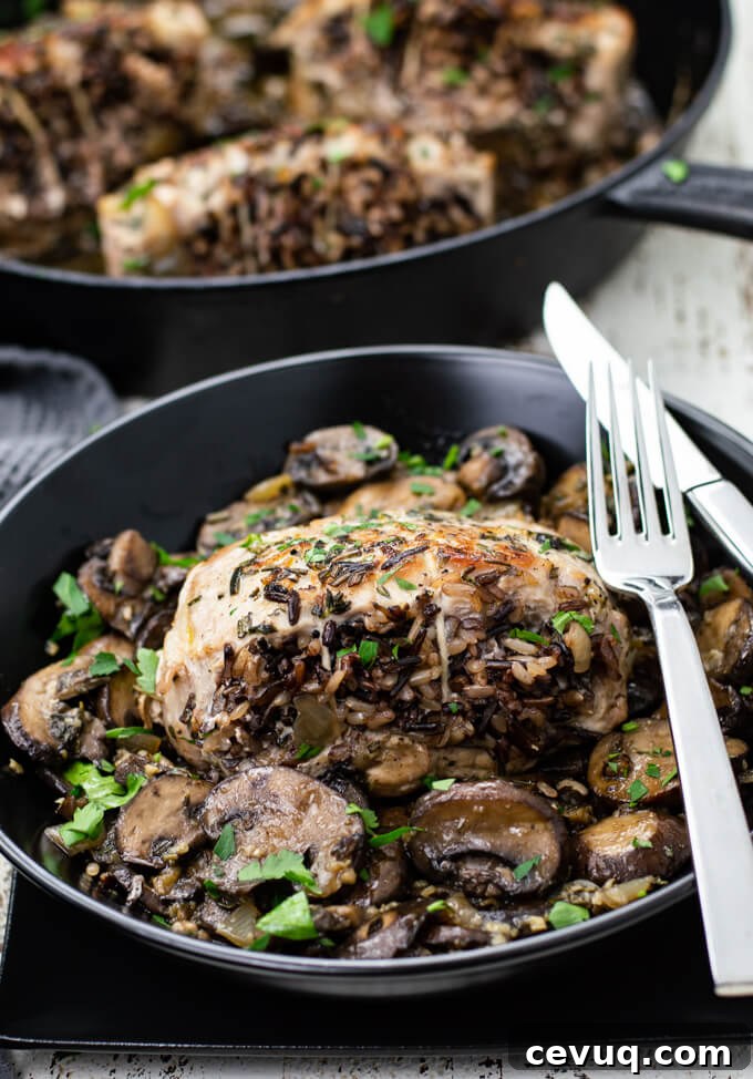 A pork chop stuffed with wild rice sits on top of sautéed mushrooms in a black dish. A fork and knife sit on the dish with a pan with more chops sit in the background.