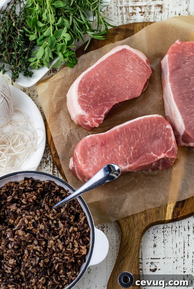 Three raw pork loin chops on a piece of parchment paper next to a b owl of wild rice, kitchen twine and fresh herbs.