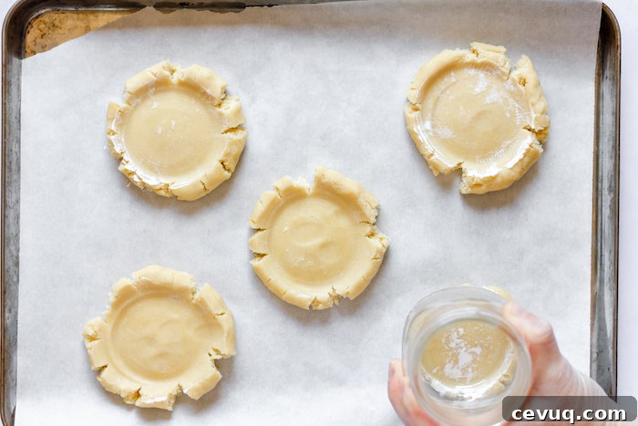 Image, taken from above, shows five sugar cookies with a hand pressing a glass to flatten them. 