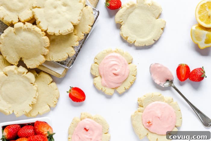 Image shows a counter top with several sugar cookies on a cooling rack and the counter. A spoon with pink frosting rests nearby along with some strawberries. 