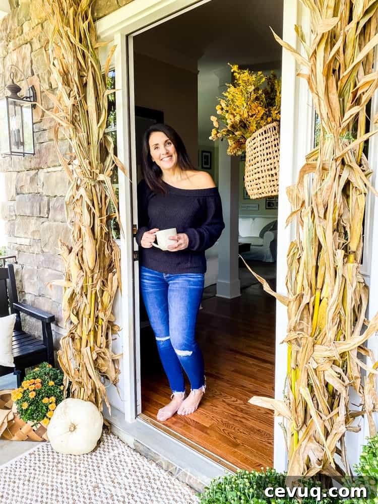 woman leaning against door frame of open front door decorated with cornstalks, flowers and pumpkins
