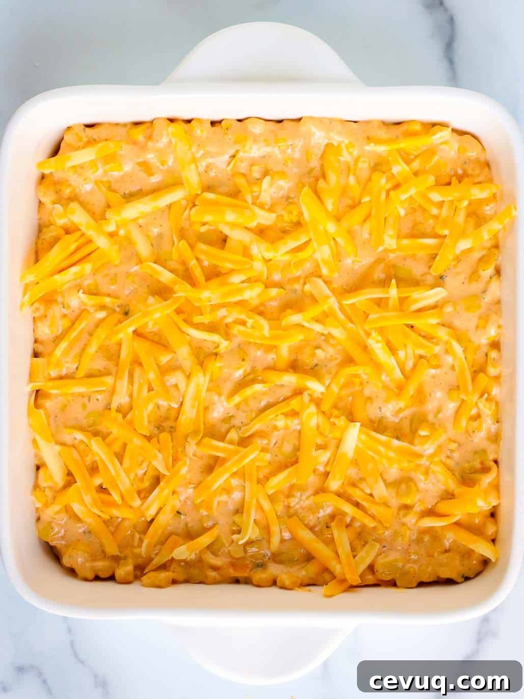 Cheesy corn casserole mixture is transferred into in an 8 by 8 inch baking dish and topped with shredded cheese.
