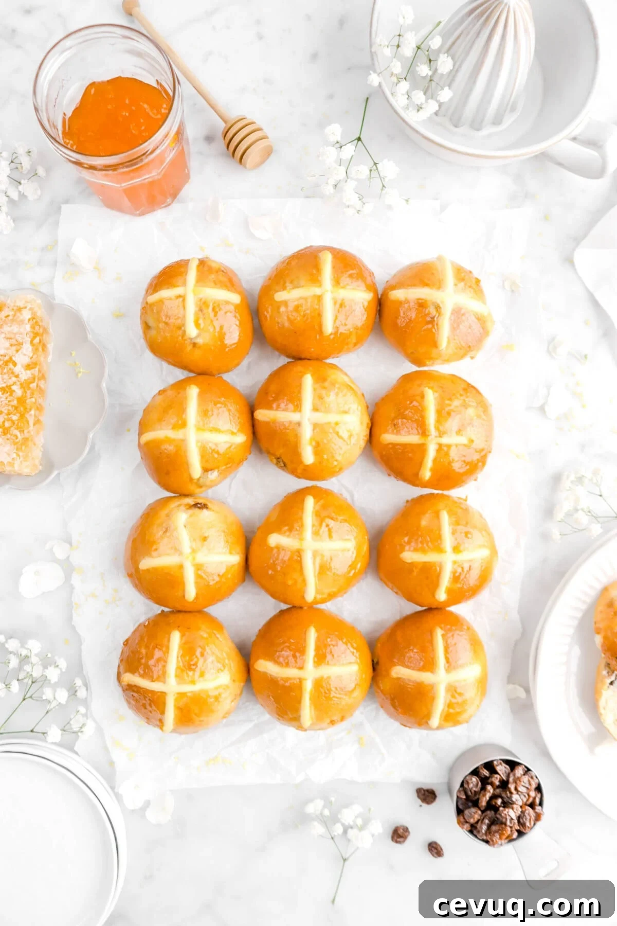 overhead shot of 12 hot cross buns on parchment paper with flowers, citrus juicer, jar of jam, wooden honey dipper, honey comb, and measuring cup of raisins around