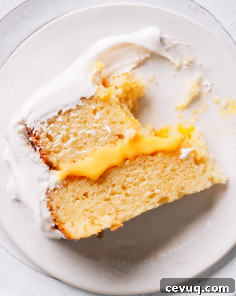 Slice of lemon curd cake on white plate with layers of lemon cake, tangy lemon curd filling, and fluffy white frosting. A portion cut with a fork is visible.