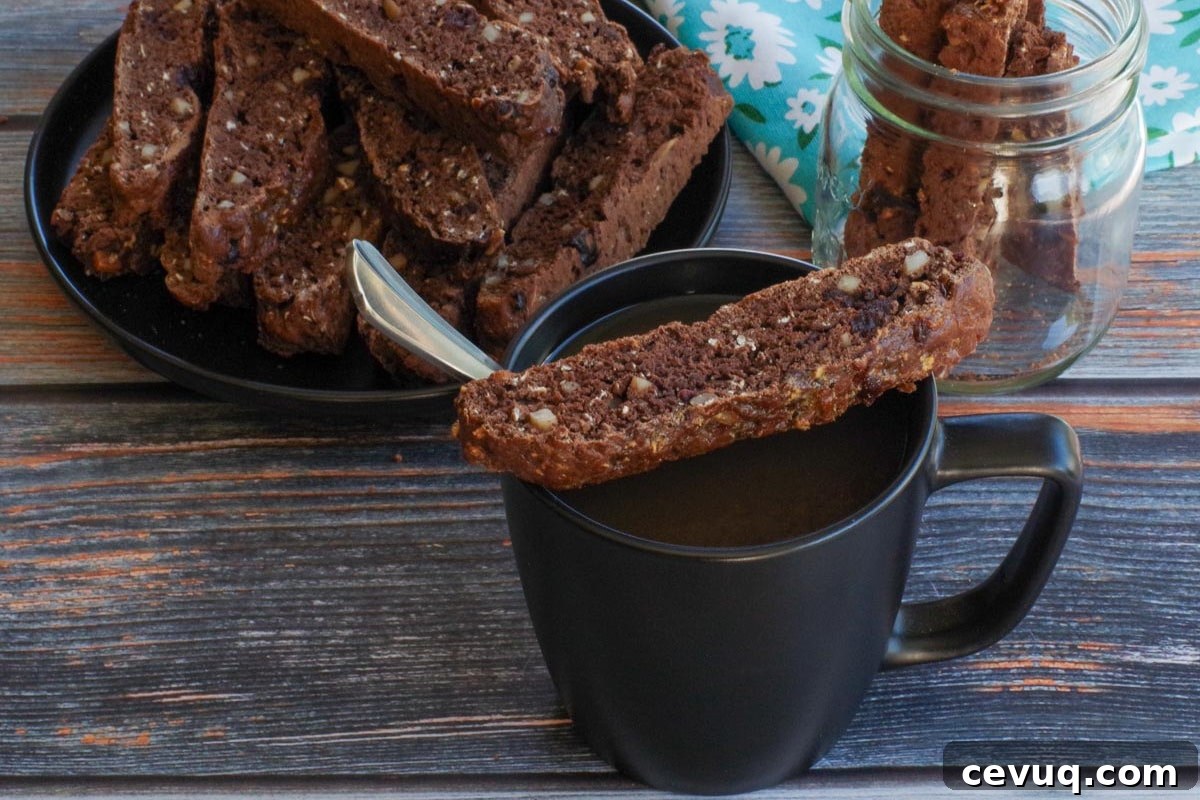 Chocolate Zucchini Biscotti Recipe: Crunchy, Moist, and Perfect for Coffee 2 zucchini biscotti resting on top of a black coffee mug with a plate of more biscotti and a jar in the background