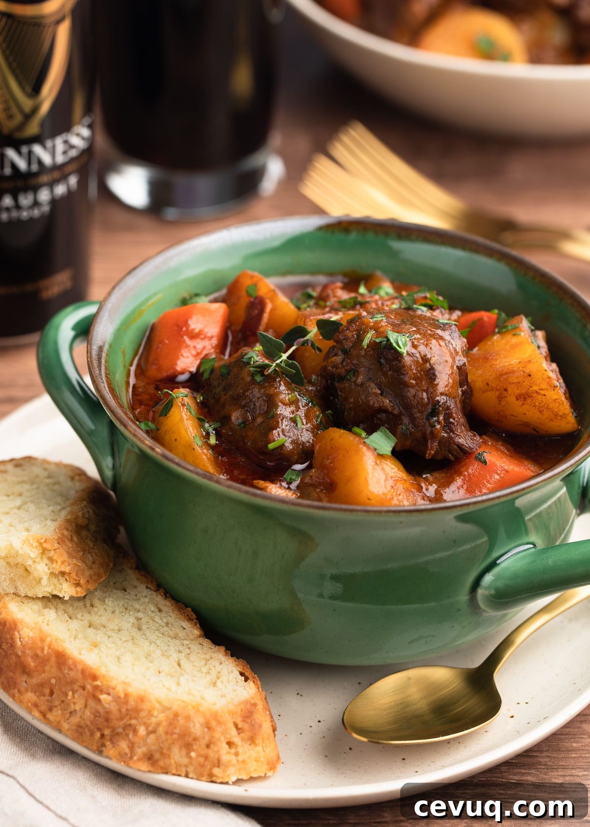 Guinness Beef Stew in a green handled bowl on top of an ivory plate with slices of Irish Soda Bread and a gold tone spoon