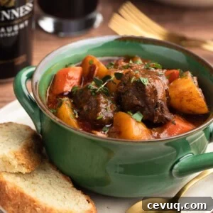 Guinness Beef Stew in a green handled bowl on top of an ivory plate with slices of Irish Soda Bread and a gold tone spoon