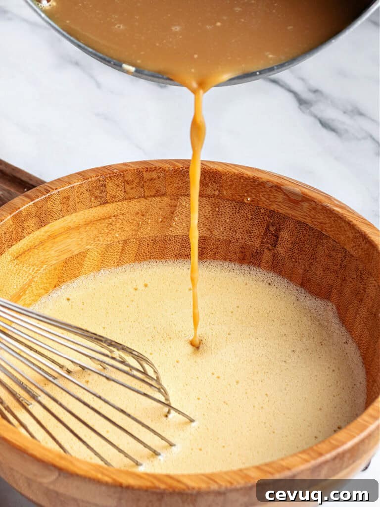 Pouring brown sugar milk into beaten egg yolks in a wooden bowl. A wire whisk.