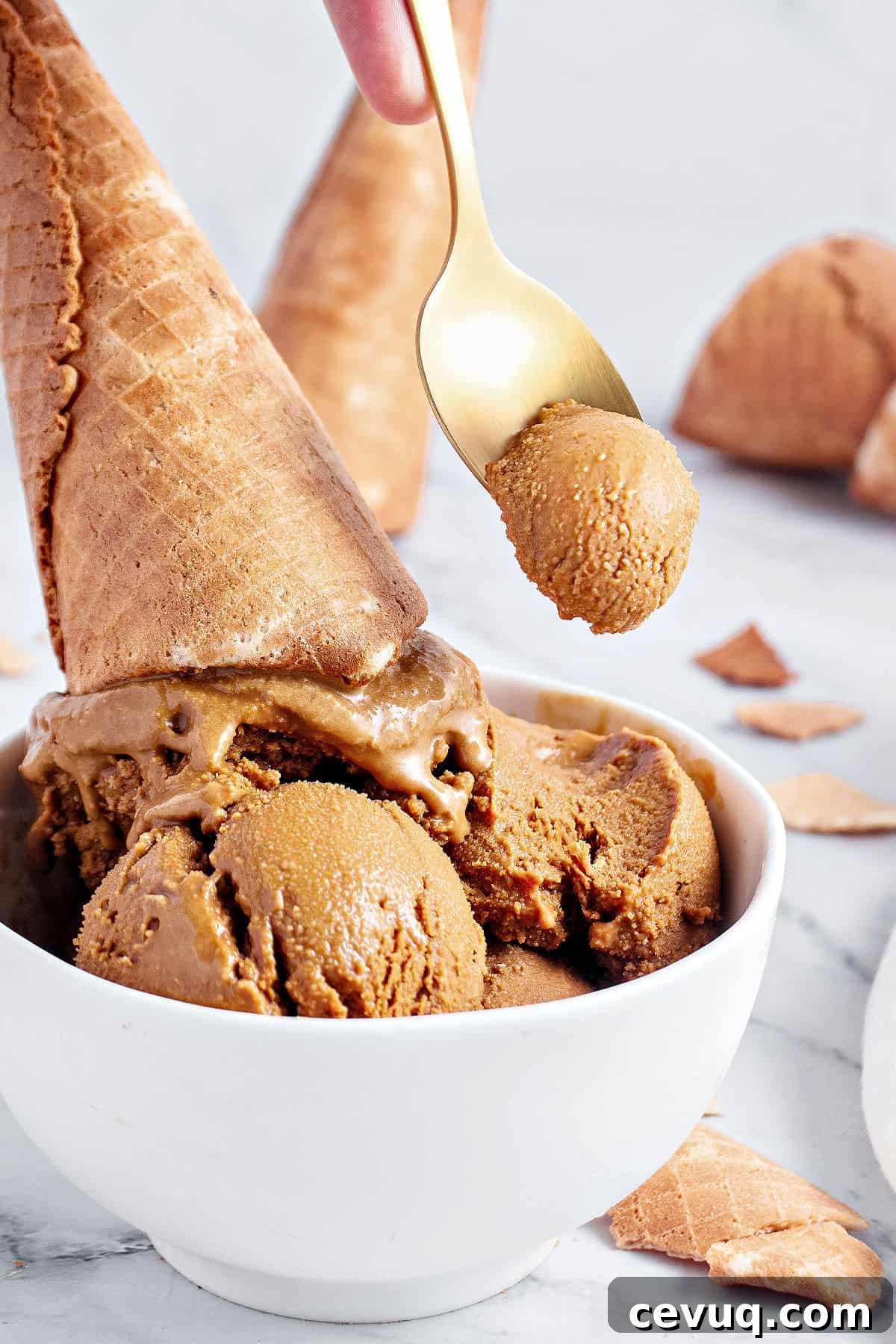 Waffle cone on scoops of brown sugar ice cream in a white bowl. Golden spoon, gray background.