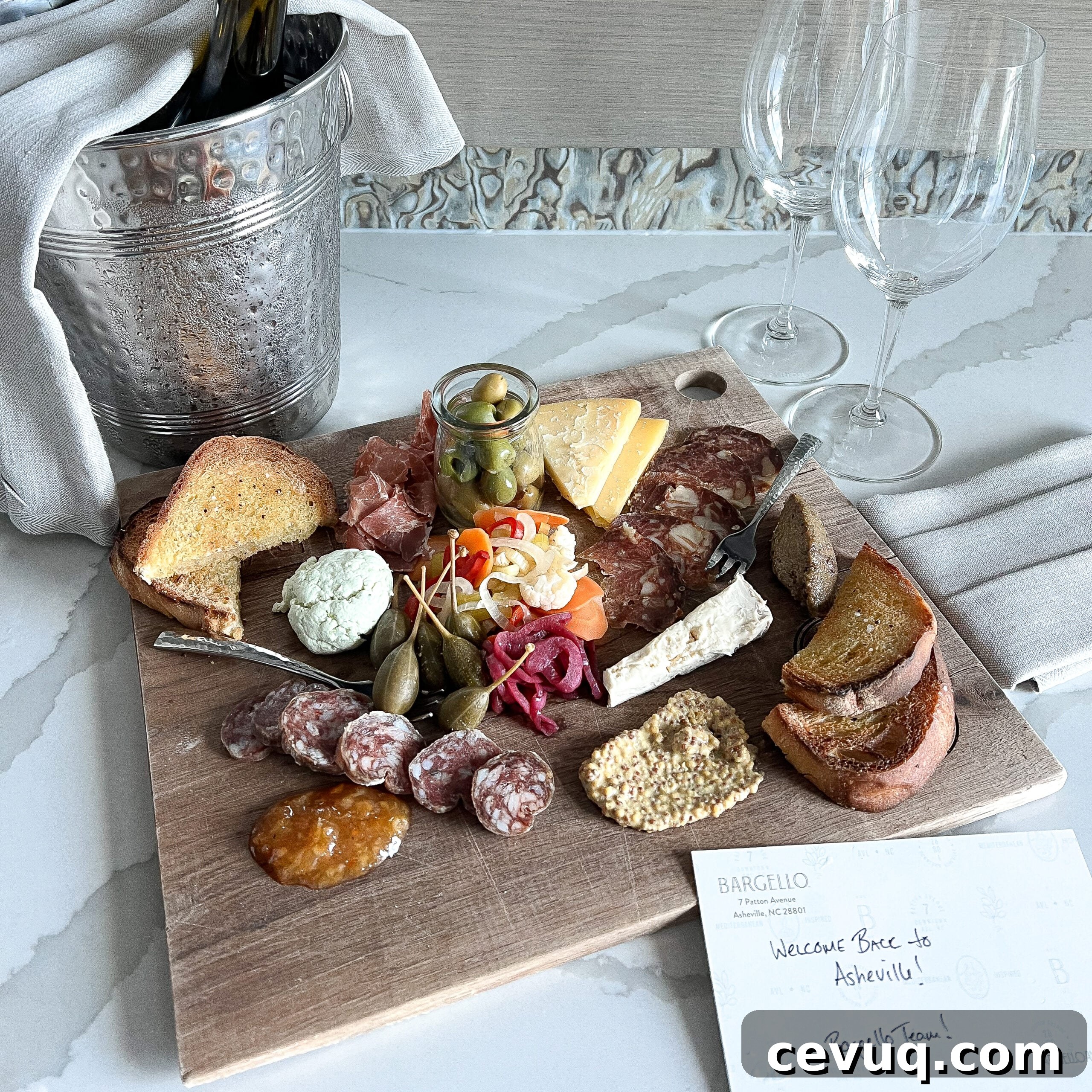 A wooden charcuterie board with cheeses, olives, pickles and bread beside wine glasses.