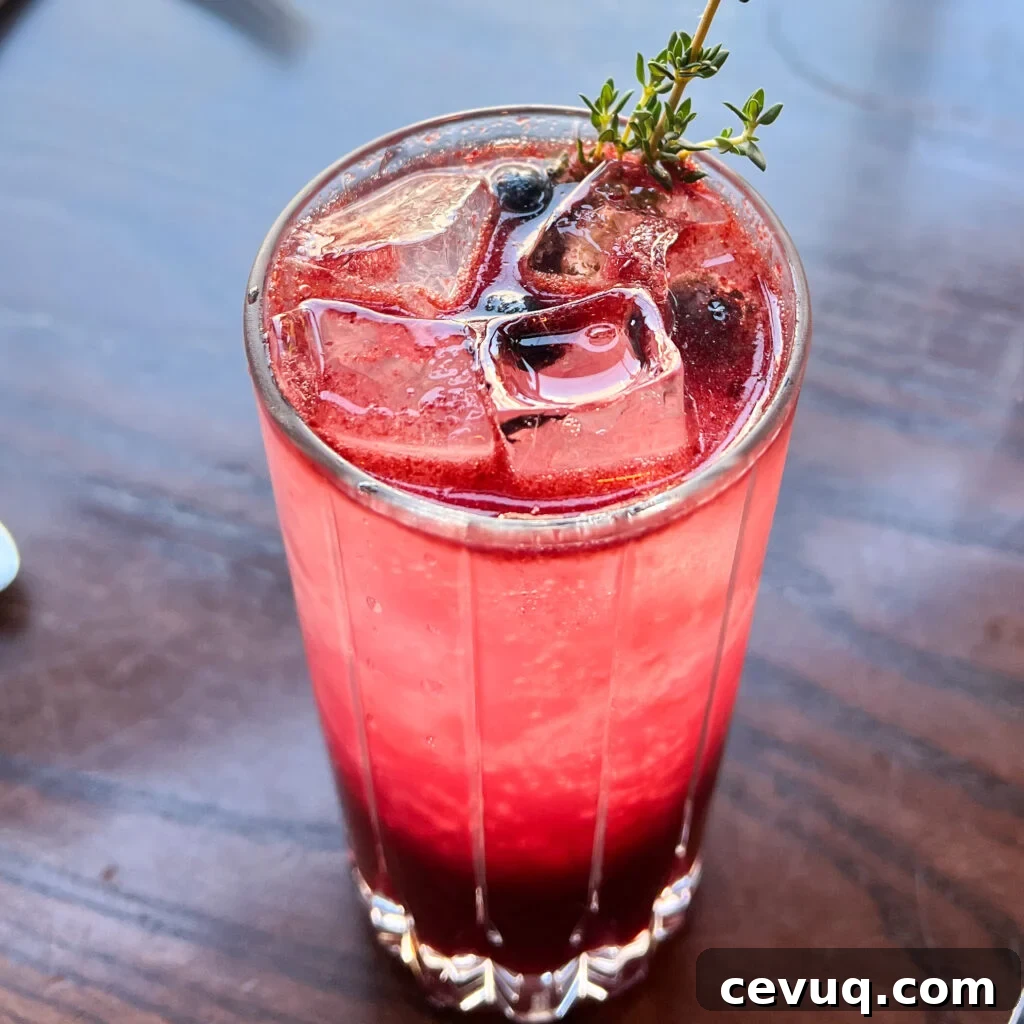 A tall glass of berry cocktail garnished with thyme on a wooden table.
