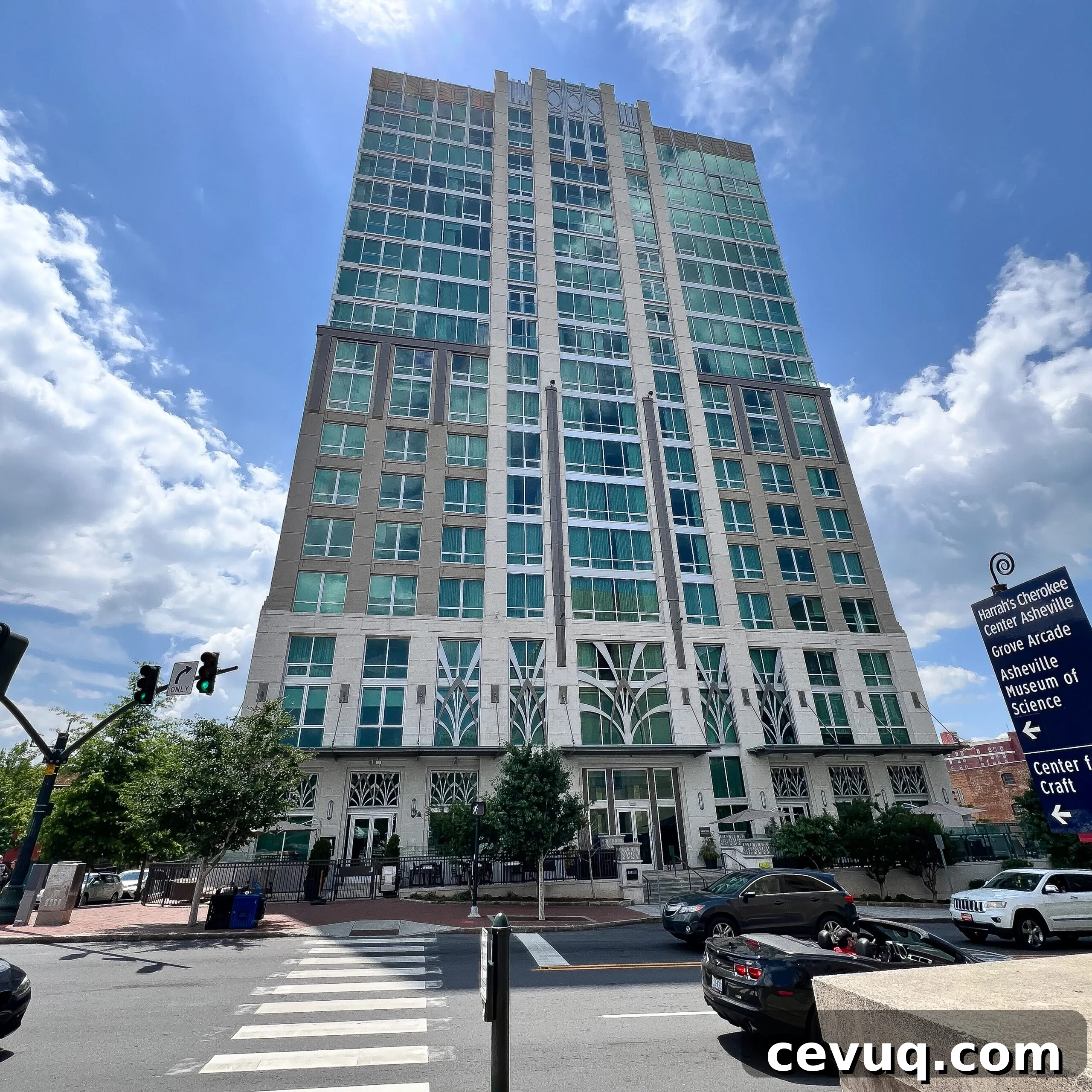 A tall, modern building with green-tinted windows stands on a city street corner under a partly cloudy sky.