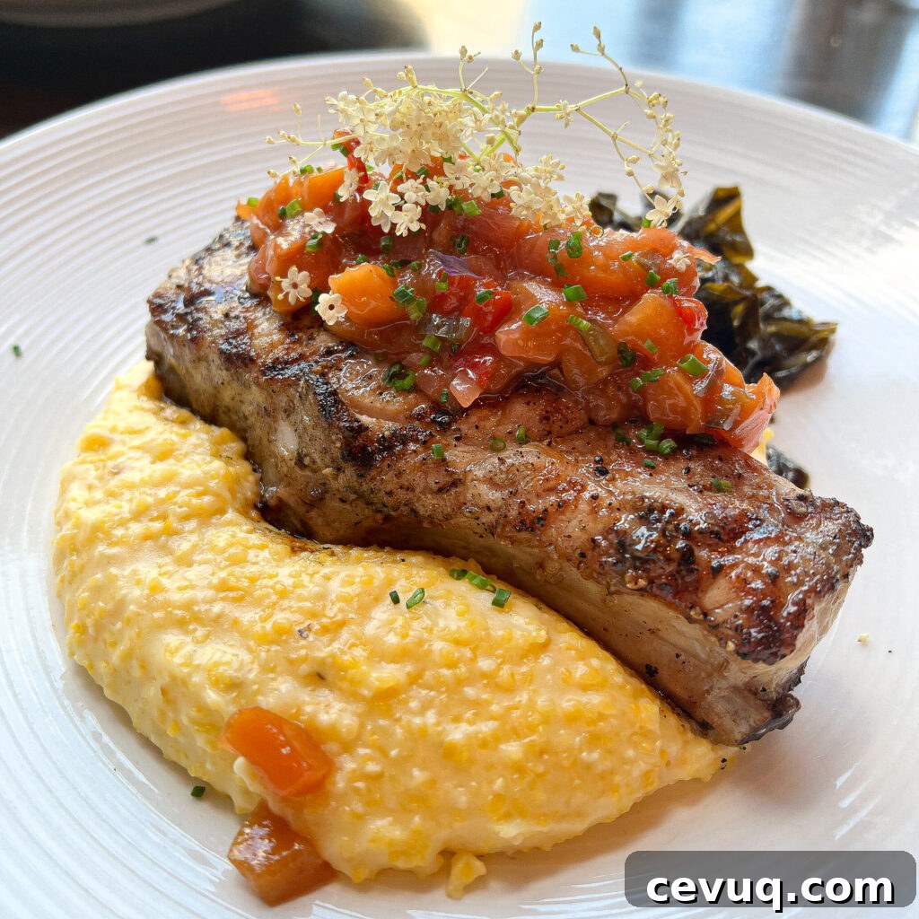 A plated pork chop served with grits and sautéed greens, garnished with edible flowers.