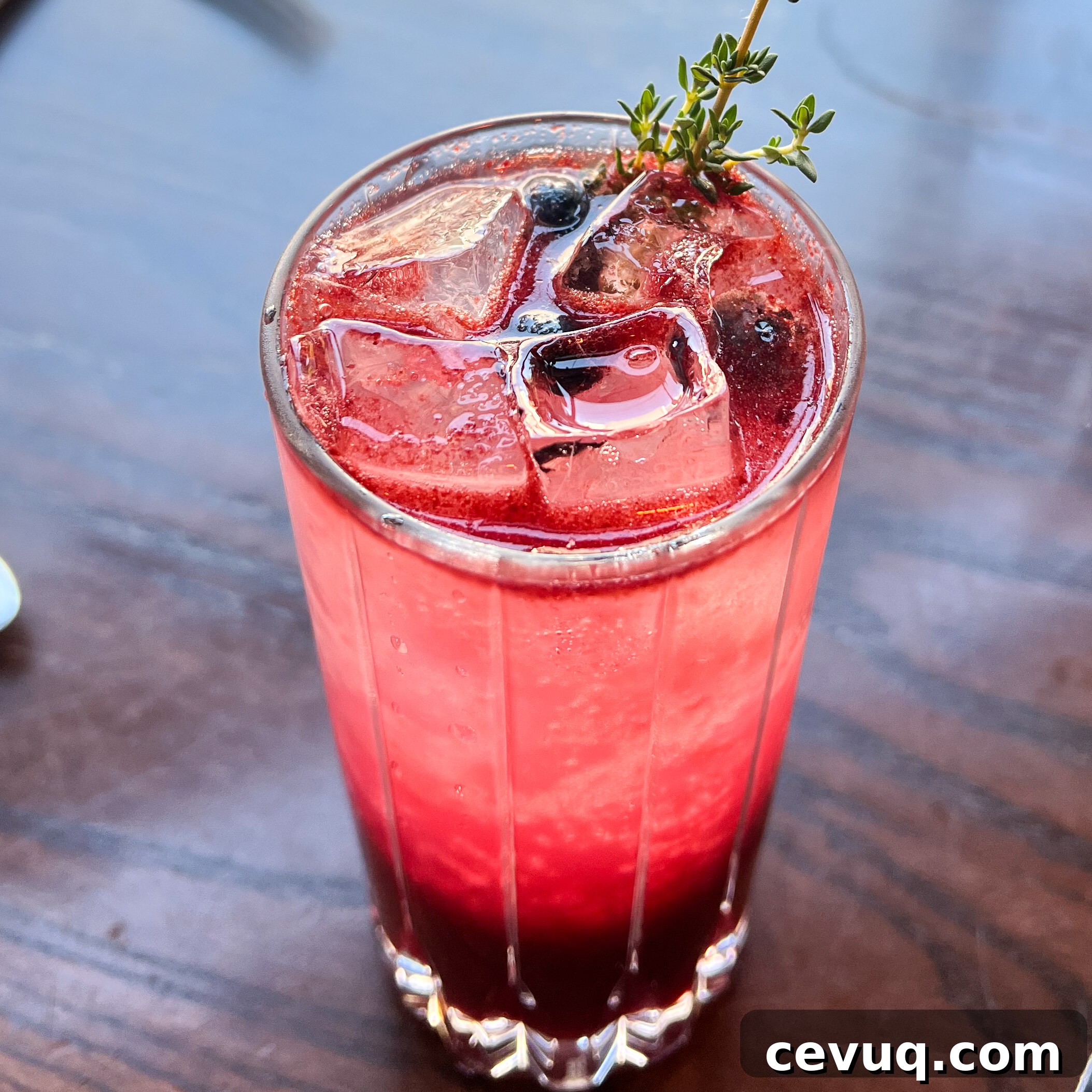A tall glass of berry cocktail garnished with thyme on a wooden table.