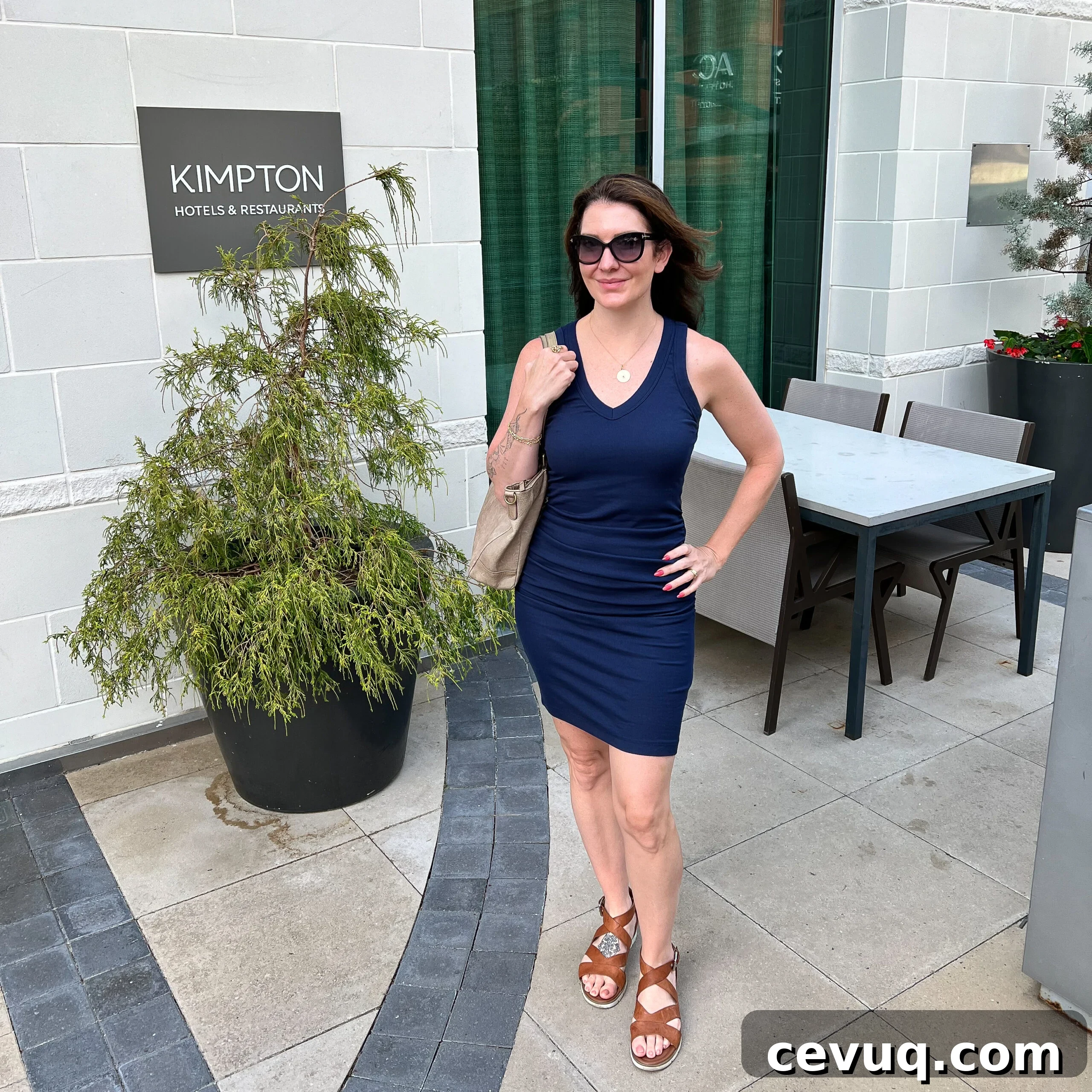 A woman in a navy blue dress poses outside a hotel near patio seating.