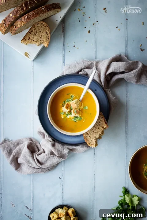 A bowl set on a blue plate over a light brown linen napkin, filled with carrot and coriander soup that's topped with croutons, soured cream and coriander. Set on a cool grey wood effect backdrop, there's also a spoon in the soup and a torn piece of bread on the side. Around the edge of the image is a sliced loaf of bread, a pot of croutons and some coriander.