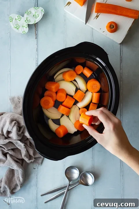 A hand placing a piece of carrot in a slow cooker that's filled with carrots and onions. There's a white chopping board with carrot on it, some stock pots, spoons and a light brown linen napkin in the background. Set on a cool grey wood effect backdrop.
