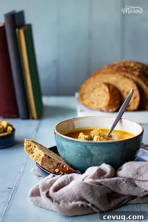 A front view of a bowl of slow cooker carrot and coriander soup with a spoon inside, topped with croutons, soured cream and coriander. Set on a light brown linen napkin over a cool grey wood effect backdrop, there's also a sliced loaf of bread, a pot of croutsons and some books in the background.