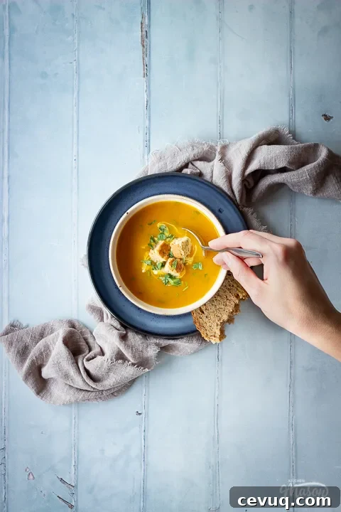 A hand reaching in with a spoon over a bowl set on a blue plate over a light brown linen napkin, filled with carrot and coriander soup that's topped with croutons, soured cream and coriander. Set on a cool grey wood effect backdrop, there's also a spoon on the side and a torn piece of bread.