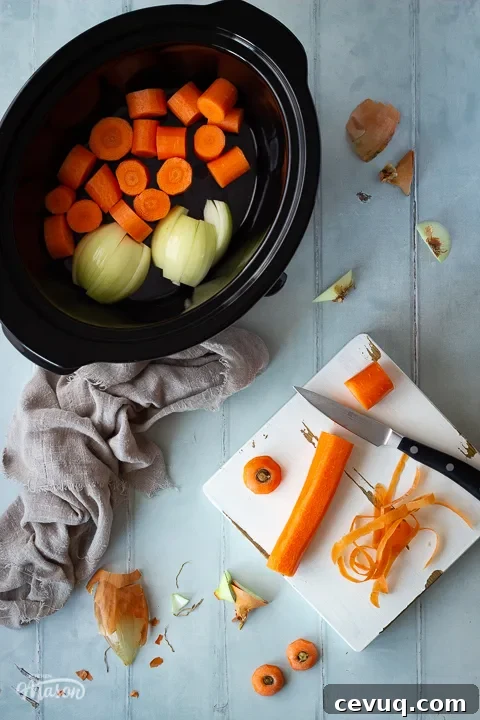 A carrot being chopped on a wooden chopping board set on a cool grey wood effect backdrop. There's also a light brown linen napkin, a slow cooker filled with vegetables and some vegetable peelings in the background.