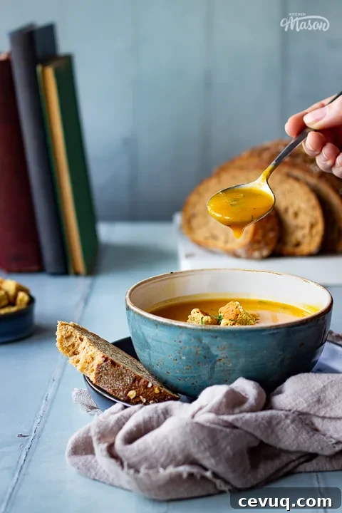 A front view of a spoon filled with soup over a bowl of slow cooker carrot and coriander soup, topped with croutons, soured cream and coriander. Set on a light brown linen napkin over a cool grey wood effect backdrop, there's also a sliced loaf of bread, a pot of croutsons and some books in the background.